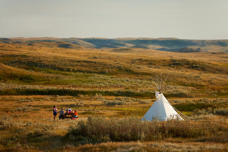 Une famille à une table de pique-nique près d'un tipi sur un champ, parc national des Prairies. Crédit Parcs Canada